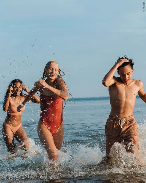 Tre giovani adulti in costume da bagno ridono e sguazzano mentre camminano nell'acqua bassa del mare in una giornata di sole.
