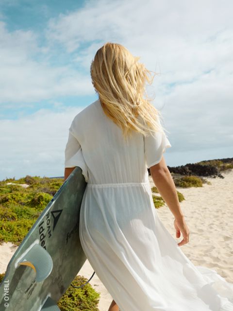 Una donna in abito bianco cammina su una spiaggia sabbiosa con una tavola da surf in mano, i suoi capelli biondi sventolano al vento.