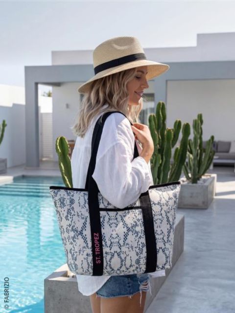 Una donna con un cappello di paglia e una camicia bianca è in piedi accanto a una piscina, tenendo una grande borsa a fantasia con la scritta "St. Tropez", con dei cactus sullo sfondo.