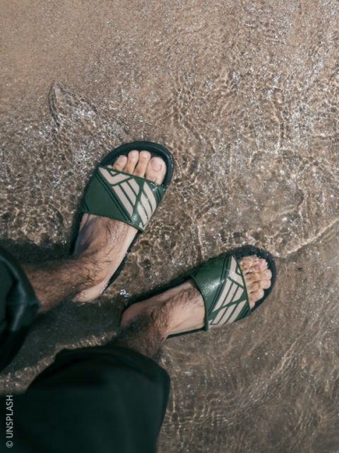 Una persona con sandali verdi sta in piedi nell'acqua bassa su una spiaggia di sabbia, con i pantaloni leggermente arrotolati.