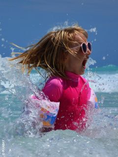 Una ragazza con occhiali da sole rosa e costume da bagno sguazza nel mare e sorride eccitata mentre l'acqua le schizza intorno in una giornata di sole.
