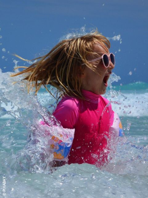 Una ragazza con occhiali da sole rosa e costume da bagno sguazza nel mare e sorride eccitata mentre l'acqua le schizza intorno in una giornata di sole.