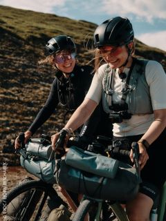 Due donne con caschi e occhiali da sole sorridono mentre vanno in bicicletta all'aperto, con borse attaccate. Sullo sfondo si vede una collina erbosa.