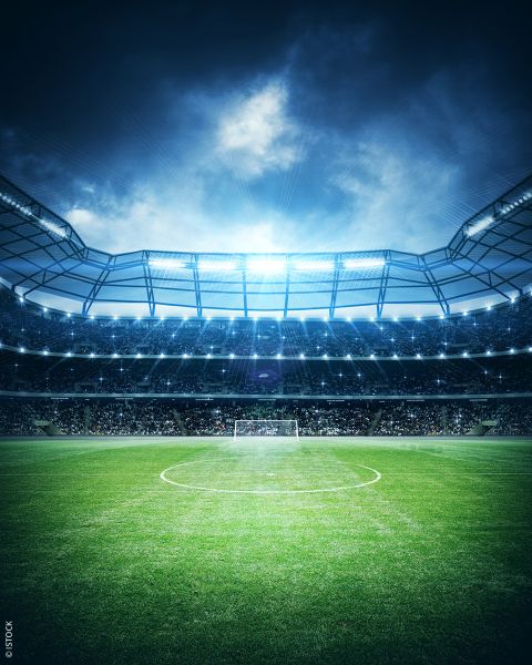 Sguardo drammatico su uno stadio di calcio illuminato con campo da gioco verde e pali della porta, pieno di tifosi esultanti sotto un cielo serale nuvoloso.