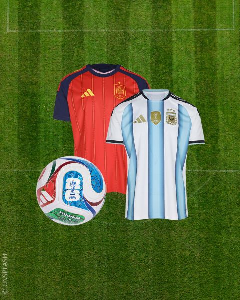 Due maglie da calcio - una rossa e una bianca con strisce blu - accanto a un pallone da calcio colorato su un campo da calcio verde.