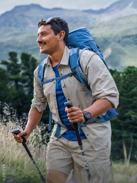 Un uomo che cammina nella natura con bastoncini da trekking, indossando uno zaino, una camicia beige e pantaloncini, e sorride sullo sfondo di alberi verdi e montagne.