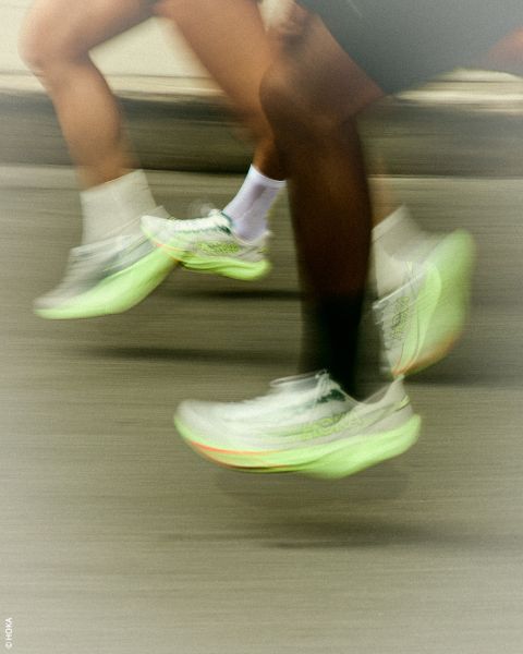 Due persone che corrono con sneaker HOKA verde neon; la sfocatura di movimento sottolinea il loro movimento veloce su una strada.