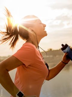 Una donna con una maglietta rosa che fa jogging all'aperto all'alba, sorride, indossa le cuffie e tiene una bottiglia d'acqua in mano, mentre il sole splende dietro di lei.