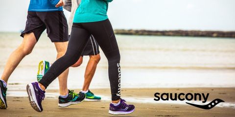 Tre persone corrono in abbigliamento sportivo lungo una spiaggia di sabbia; nell'angolo in basso a destra è visibile il logo Saucony.