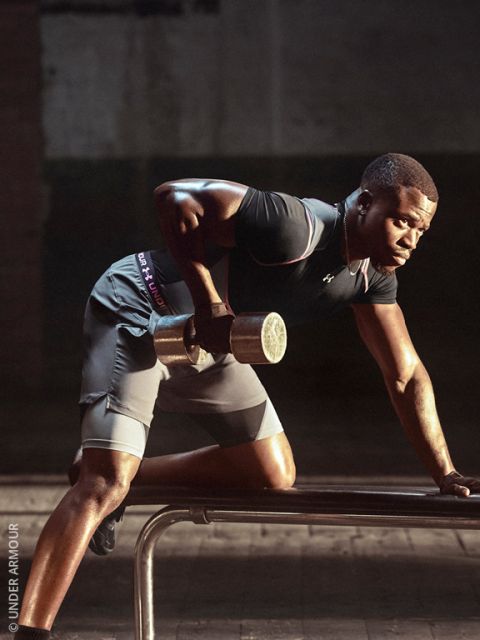Un uomo in abbigliamento sportivo solleva un bilanciere appoggiandosi a una panca; si allena concentrato in una palestra scarsamente illuminata.