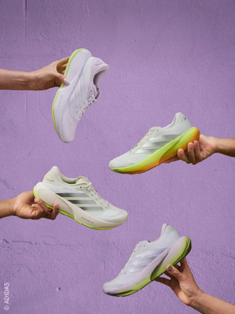 Quattro mani che tengono scarpe sportive bianche con suole verdi e gialle davanti a una parete color lavanda.