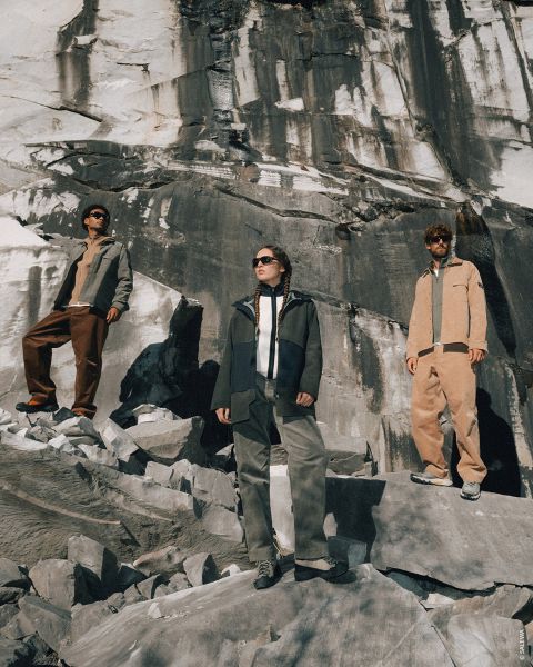 Tre persone in abbigliamento da esterno si trovano su un terreno roccioso, con un grande muro di pietra dietro di loro, e guardano in lontananza sotto un sole splendente.