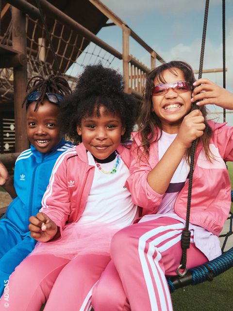 Tre bambini sorridenti in abiti Adidas sono seduti fuori su un'altalena - due di loro in tute rosa e uno in una blu - e si godono la giornata di sole.