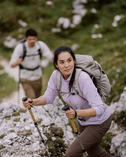 Due immagini su sfondo di legno mostrano una modella e un modello in abbigliamento da trekking outdoor. La modella indossa un outfit rosa e verde, il modello un outfit verde e tiene in mano dei bastoncini da trekking. Testo: "TIPO OUTDOOR ESCURSIONISTA ATTIVO".