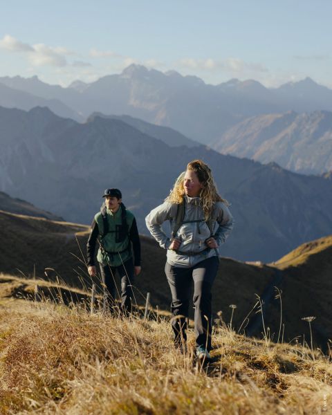 Due persone sono visibili in foto separate. La foto a sinistra mostra una persona in abbigliamento da trekking con bastoncini da trekking, quella a destra un'altra persona con zaino e giacca. Lo sfondo è una struttura in legno con muschio. Il testo recita "Tipo da outdoor Escursionista per piacere".