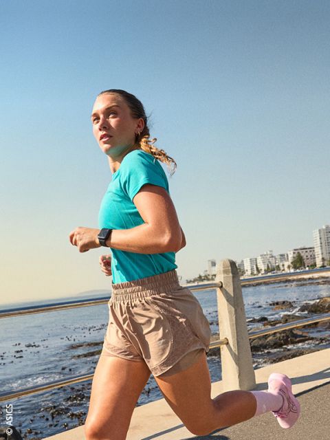 Una donna in abbigliamento sportivo fa jogging lungo il mare in una giornata di sole, sullo sfondo si vedono gli edifici della città.