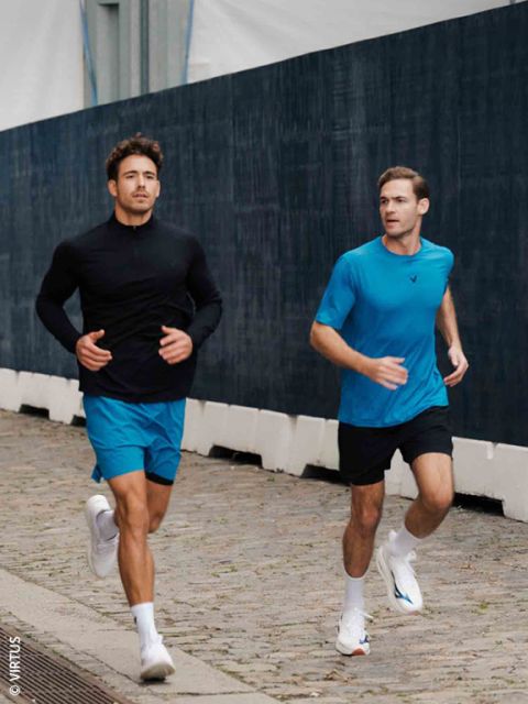 Due uomini in abbigliamento sportivo fanno jogging su un sentiero di ciottoli accanto a una recinzione scura; entrambi indossano pantaloncini blu e scarpe da ginnastica bianche.