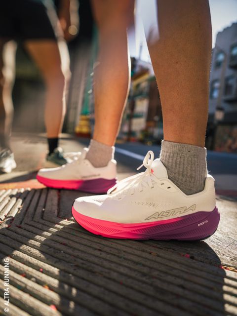 Una persona con scarpe da corsa bianche e rosa sta su un marciapiede in città; la luce del sole fa risaltare le scarpe e le gambe, mentre lo sfondo urbano appare sfocato.