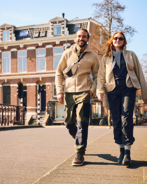 Un uomo e una donna sorridenti camminano all'aperto in una giornata di sole, vestiti con giacche e stivali casual, tenendosi per mano, con un grande edificio in mattoni sullo sfondo.
