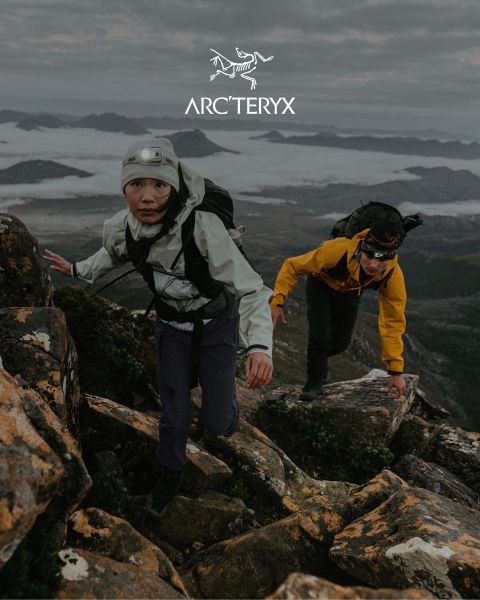 Due persone fanno escursionismo su un sentiero di montagna roccioso. Il cielo è coperto, sullo sfondo si vede una catena montuosa nebbiosa. Entrambi gli escursionisti indossano attrezzatura da outdoor. Sulla destra c'è un piccolo stagno. In alto si vede il logo Arc'teryx.