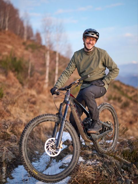 Una persona con casco e giacca verde sorride mentre posa con una mountain bike su un sentiero roccioso e innevato con uno sfondo di alberi.