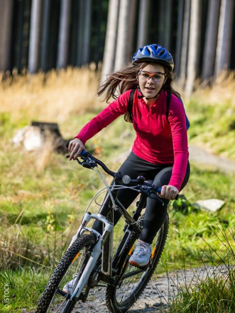 Una donna in maglietta rossa e con il casco va in mountain bike su un sentiero nel bosco sotto la luce del sole.