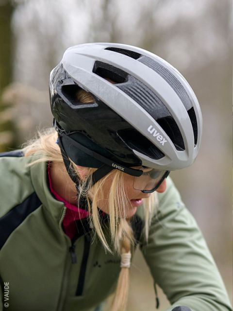 Un uomo con giacca e guanti verdi esamina un casco da bicicletta nero ed è seduto davanti a una parete di legno. Indossa una fascia nera con scritte bianche.