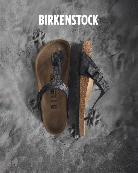 Una persona con sandali Birkenstock beige è in piedi accanto a un gioco d'acqua in un lussureggiante paesaggio di giardino. L'area è circondata dal verde e la parola "BIRKENSTOCK" è ben visibile sopra l'immagine.
