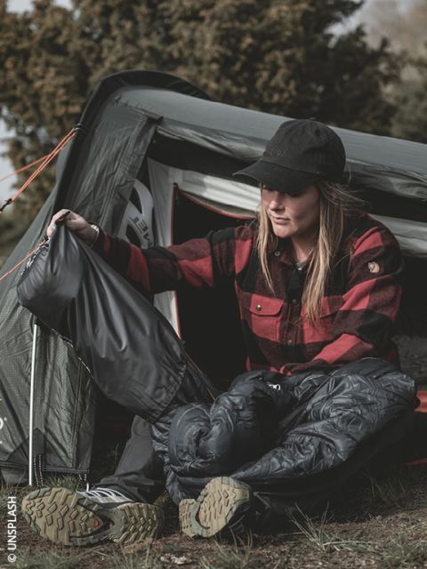 Una donna con una camicia a quadri rossa è seduta davanti a una tenda, si sta mettendo i pantaloni antipioggia e gli scarponi da trekking, preparandosi per attività all'aperto.