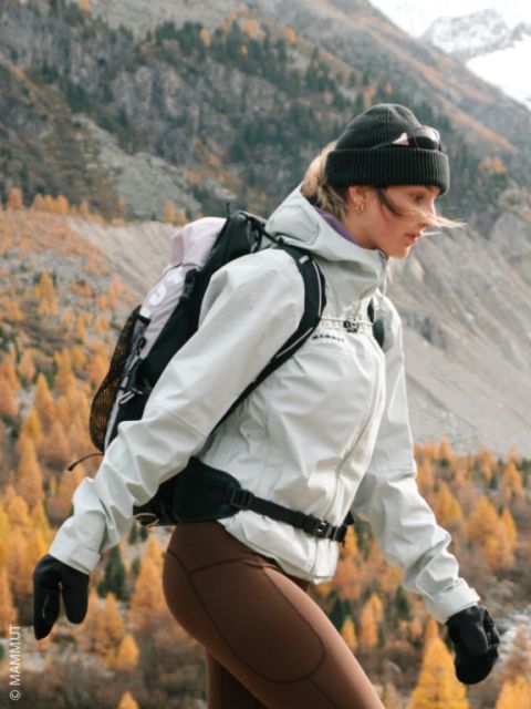 Una donna fa un'escursione in autunno nella natura selvaggia, vestita con una giacca, un cappello, guanti e uno zaino. Sullo sfondo si vedono montagne e alberi d'arancio.