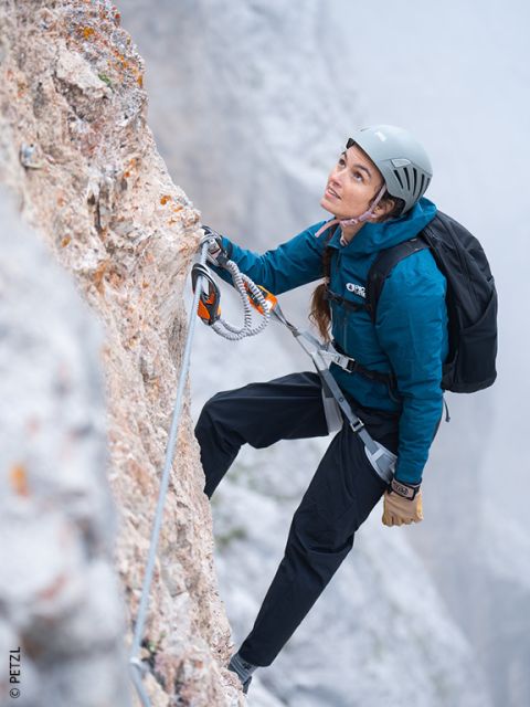 Una donna in attrezzatura da arrampicata sale su una scogliera rocciosa, assicurata da una corda di sicurezza, guardando in alto con concentrazione. Sullo sfondo c'è nebbia.