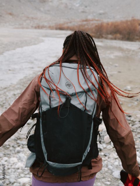 Una persona con i capelli intrecciati e un grande zaino cammina su un terreno roccioso vicino a un fiume, con la schiena rivolta verso la telecamera.