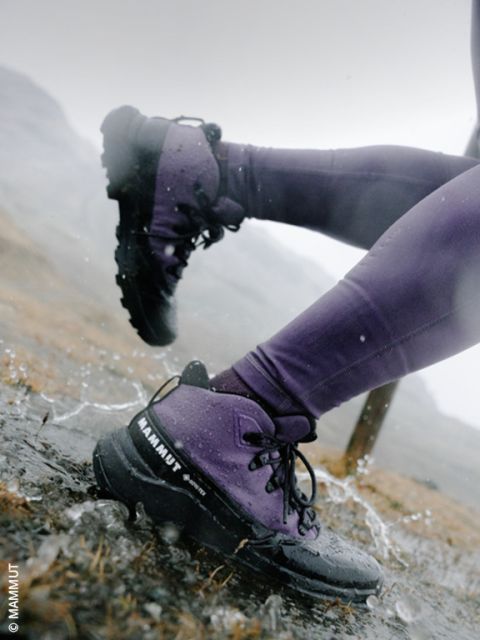 Primo piano delle gambe di una persona con pantaloni viola e scarponi da trekking neri che guada una pozzanghera su un sentiero roccioso bagnato dalla pioggia.