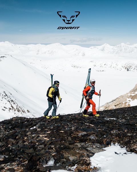 Due persone corrono su un sentiero di montagna con una città e un fiume sullo sfondo. Uno indossa una maglietta nera e pantaloncini, l'altro una canotta blu e pantaloncini. Il logo Dynafit si trova nella parte superiore dell'immagine.