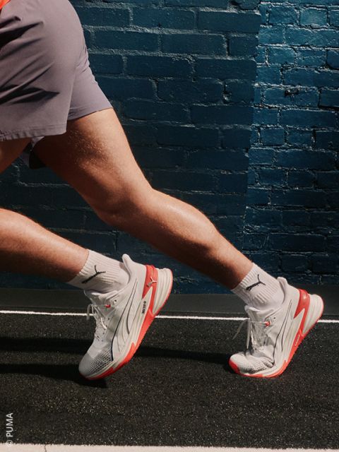 Una donna in tuta blu e bianca è seduta a terra e posa con un ginocchio sollevato. Indossa calzini bianchi e scarpe da ginnastica argentate Under Armour e sorride leggermente davanti a uno sfondo chiaro e semplice.