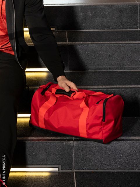 Una persona in pantaloni da jogging beige chiaro, scarpe da ginnastica nere e top nero tiene in mano un borsone nero lucido. Si trova su assi di legno scuro accanto a un vecchio faro e valigie impilate.