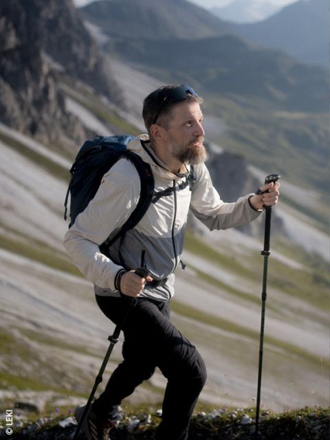 Un uomo con la barba cammina in salita su un sentiero di montagna roccioso, usando bastoncini da trekking e portando uno zaino; sullo sfondo si vedono le montagne.