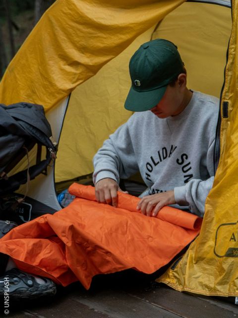 Una persona con un cappello verde è seduta in una tenda gialla e piega un materassino arancione mentre si accampa all'aperto.