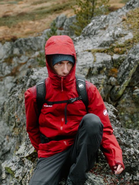 Una persona con una felpa rossa con cappuccio e uno zaino è seduta all'aperto su delle rocce, con alberi e terreno roccioso visibili sullo sfondo.