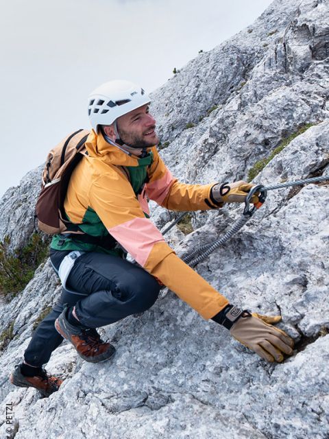 Un uomo con attrezzatura da arrampicata sale su un ripido pendio roccioso, tenendosi a una corda di sicurezza con un moschettone.