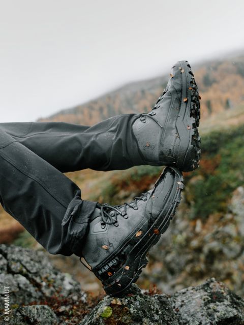 Una persona con scarponi da trekking neri, con fango e foglie attaccati, appoggia i piedi su delle rocce all'aperto, con un paesaggio montano sfocato sullo sfondo.