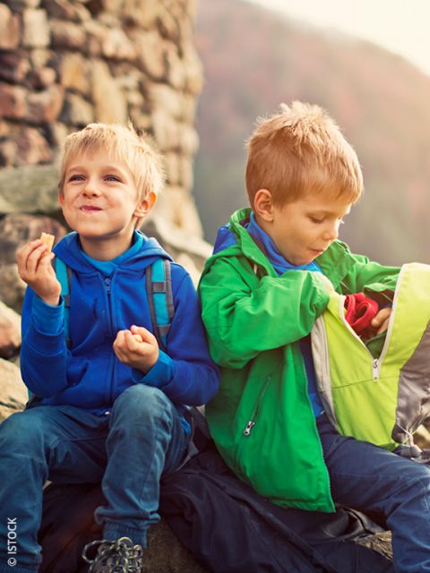 Due bambini piccoli sono seduti all'aperto sulle rocce. Uno sta mangiando uno spuntino e sorride, mentre l'altro sta frugando in uno zaino verde. Entrambi indossano giacche colorate.