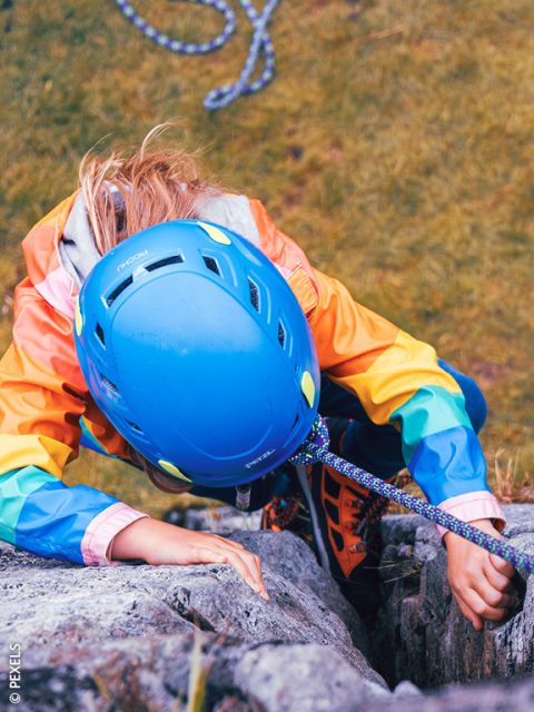 Una persona con un casco blu e una giacca colorata si arrampica su una parete rocciosa, assicurata da una corda. Vista dall'alto, sotto si vede un terreno erboso.