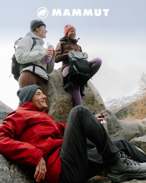 Due escursionisti con attrezzatura da outdoor attraversano un terreno roccioso sotto un cielo nuvoloso, con una grande montagna sullo sfondo. Entrambi indossano abbigliamento Mammut e il logo del marchio è visibile nella parte superiore dell'immagine.
