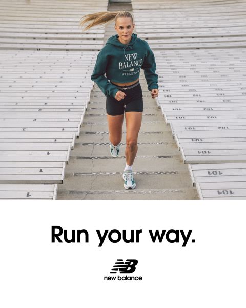 Due persone fanno jogging all'aperto, una con una giacca blu e leggings neri, l'altra con una giacca nera e leggings color castagna. Entrambe indossano scarpe da ginnastica bianche. Corrono accanto a un edificio moderno con pannelli di vetro. In basso si vede il logo New Balance.