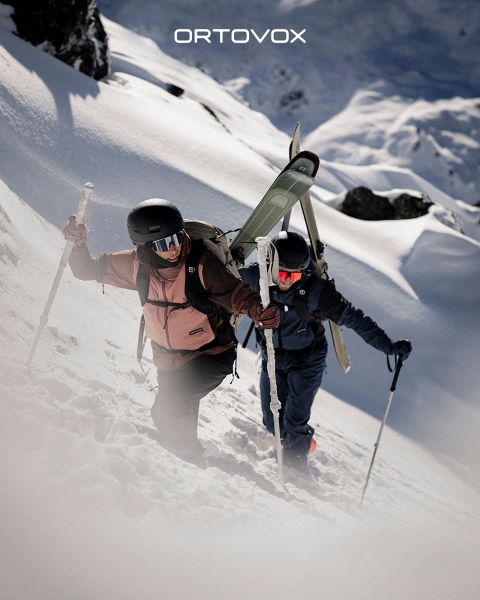 Tre persone con abbigliamento da outdoor sono sedute su un terreno roccioso in una regione montuosa, sorridendo e chiacchierando. Il sole tramonta dietro le cime. Uno zaino e un thermos sono nelle vicinanze. Il logo di Ortovox si trova in alto a destra.