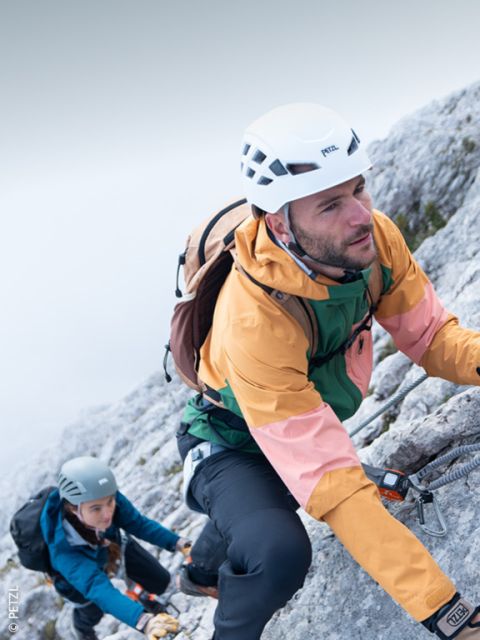 Un alpinista con casco, occhiali da sole e attrezzatura da arrampicata scala un pendio roccioso con cime innevate sullo sfondo sotto un cielo limpido.