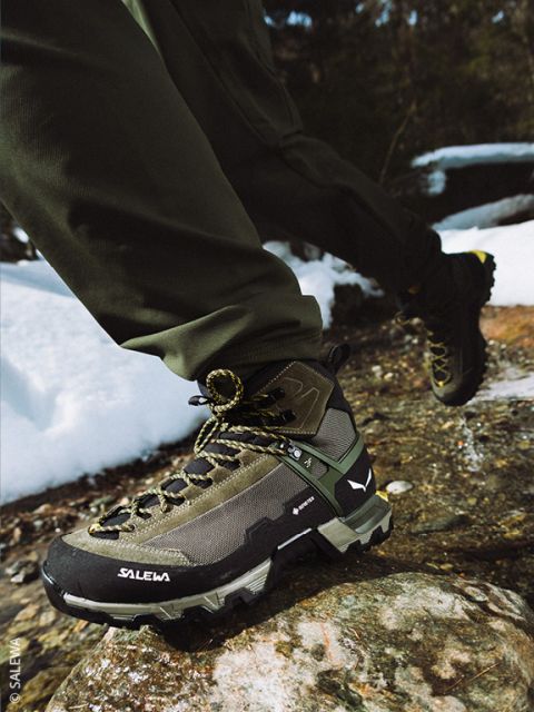 Una persona in abbigliamento da outdoor stringe i lacci di uno scarpone da trekking nero mentre si trova su una superficie rocciosa, con una grande formazione rocciosa e un cielo blu sullo sfondo.