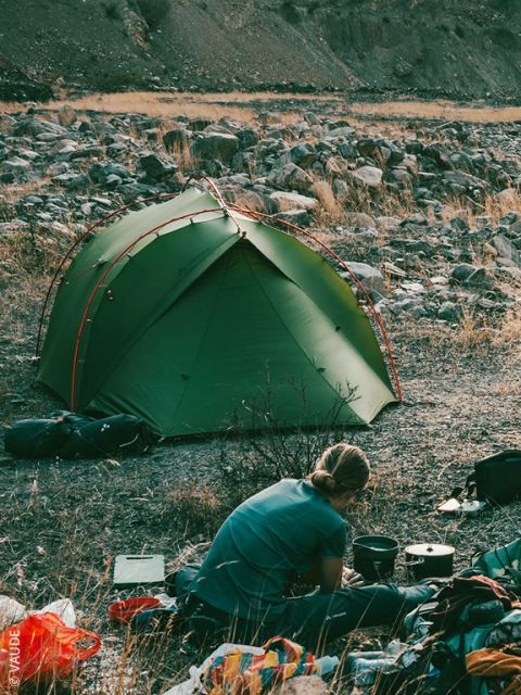 Una persona con giacca e cappello rossi prepara del cibo all'aperto, accovacciata nell'erba, vicino all'attrezzatura da campeggio, vista attraverso la porta aperta di una tenda con montagne e alberi sullo sfondo.