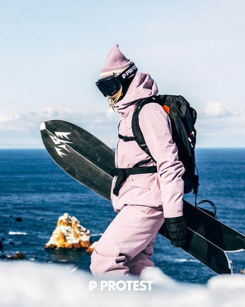 Una persona con una tuta da neve rosa e un cappello, che tiene in mano uno snowboard, si trova in riva al mare, con formazioni rocciose sullo sfondo e la parola "PROTEST" in basso.